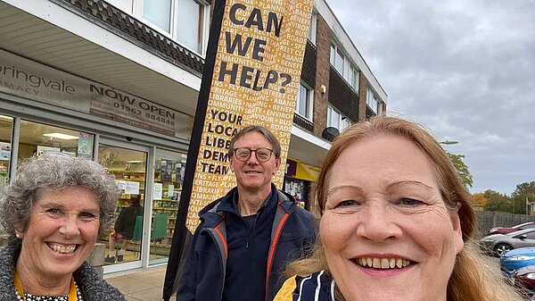 Three Liberal Democrat councillors in front of a banner saying 'Can We Help?'